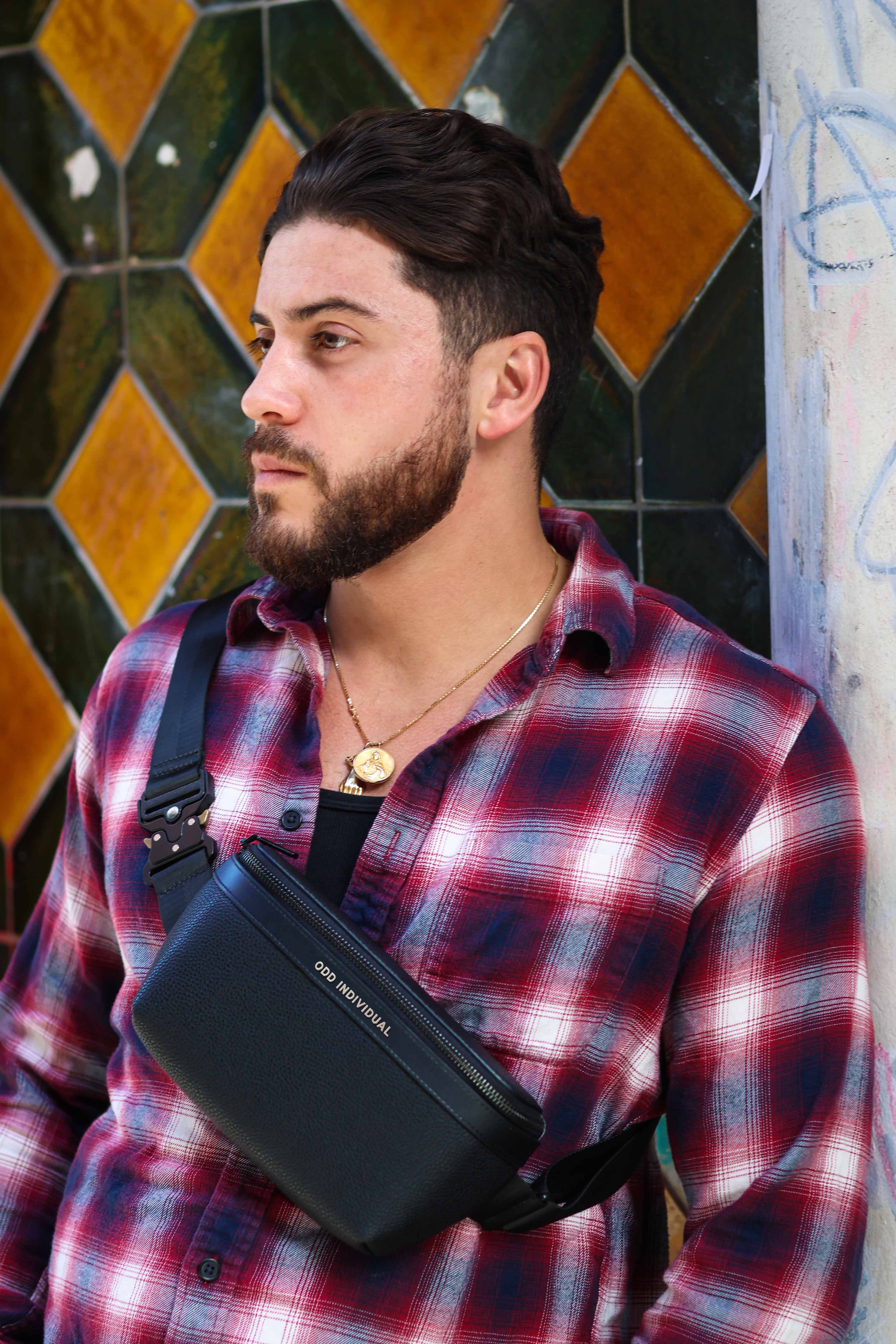 Man wearing a red plaid shirt with a black pebble leather sling bag over his shoulder against a colorful tiled wall.