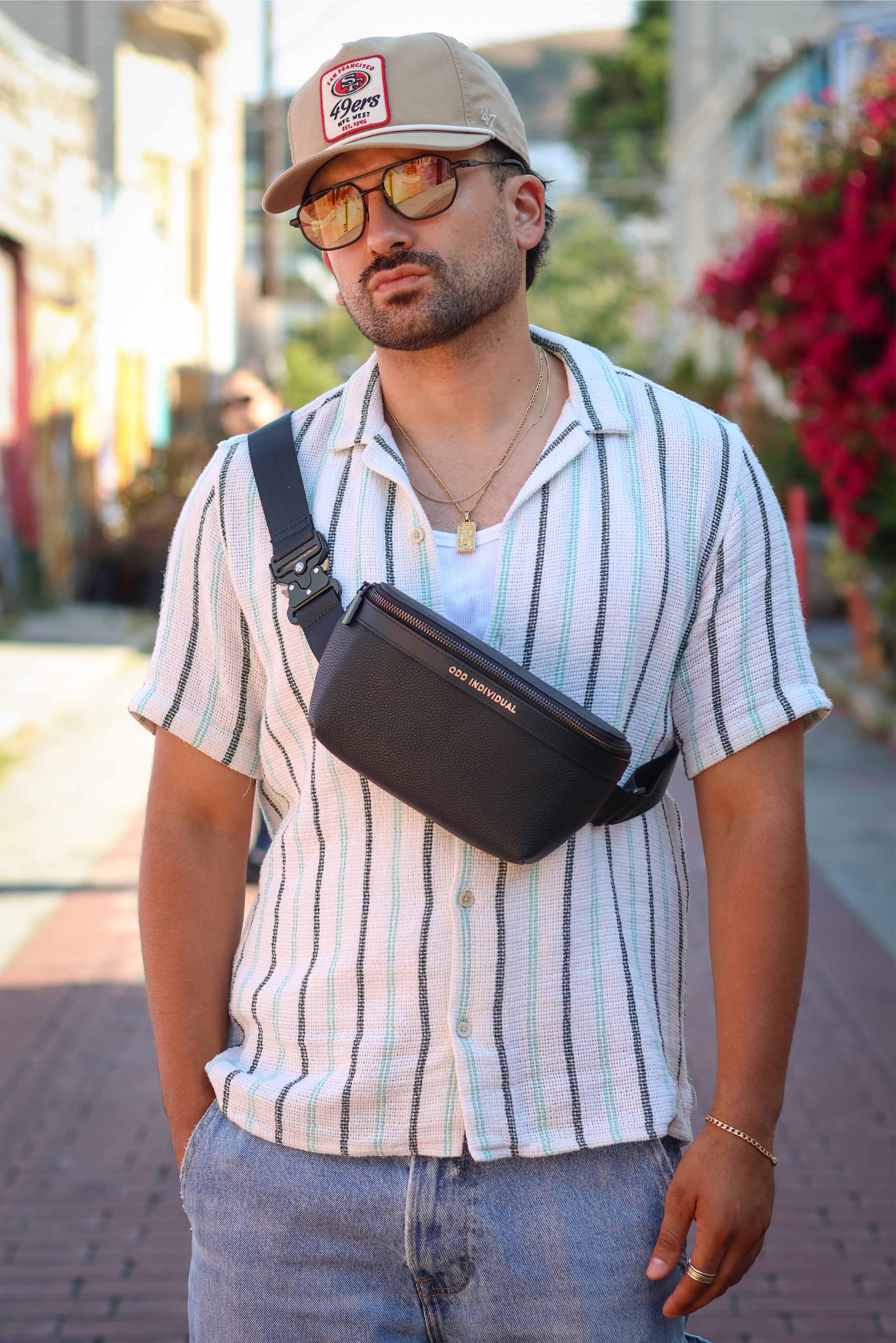 Man wearing a striped shirt, sunglasses, and a cap with a black leather sling bag/black leather belt bag/black leather crossbody bag/black leather waist bag/black leather travel pouch with brand name 'ODD INDIVIDUAL' on a street.