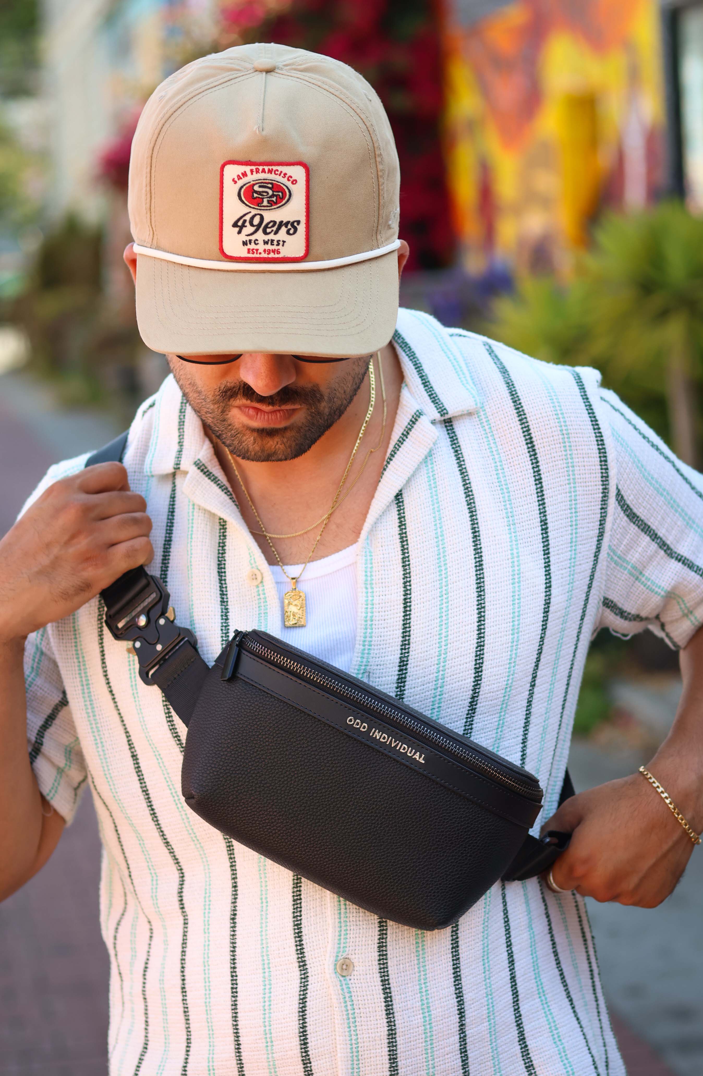 Man wearing a cap and striped shirt, wearing and holding a black leather sling bag/black leather belt bag/black leather crossbody bag/black leather waist bag/black leather travel pouch with brand name 'ODD INDIVIDUAL' with a blurred colorful background