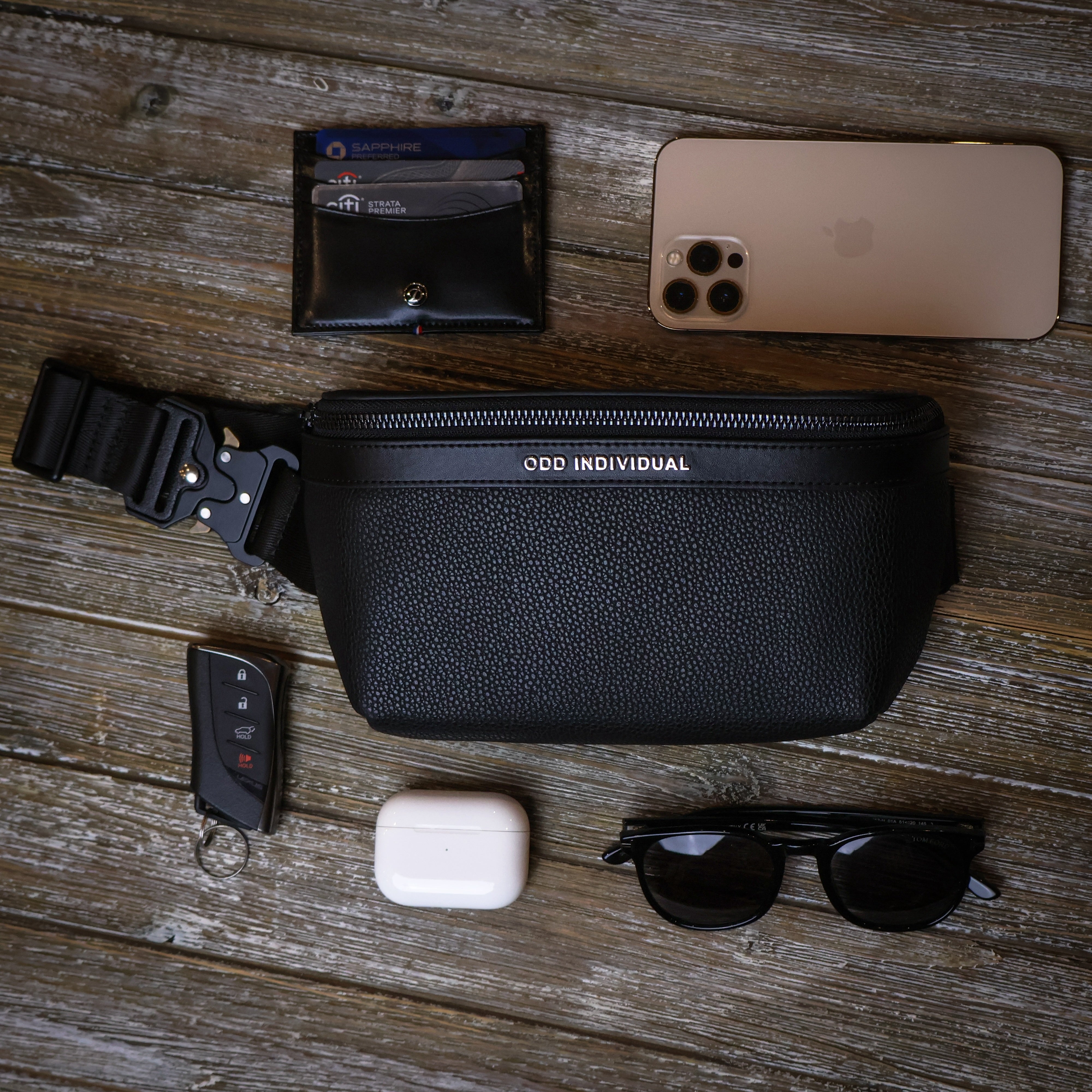 Men's sling bag with black pebble leather with phone, wallet, sunglasses, Airpods, and car keys on a wooden surface
