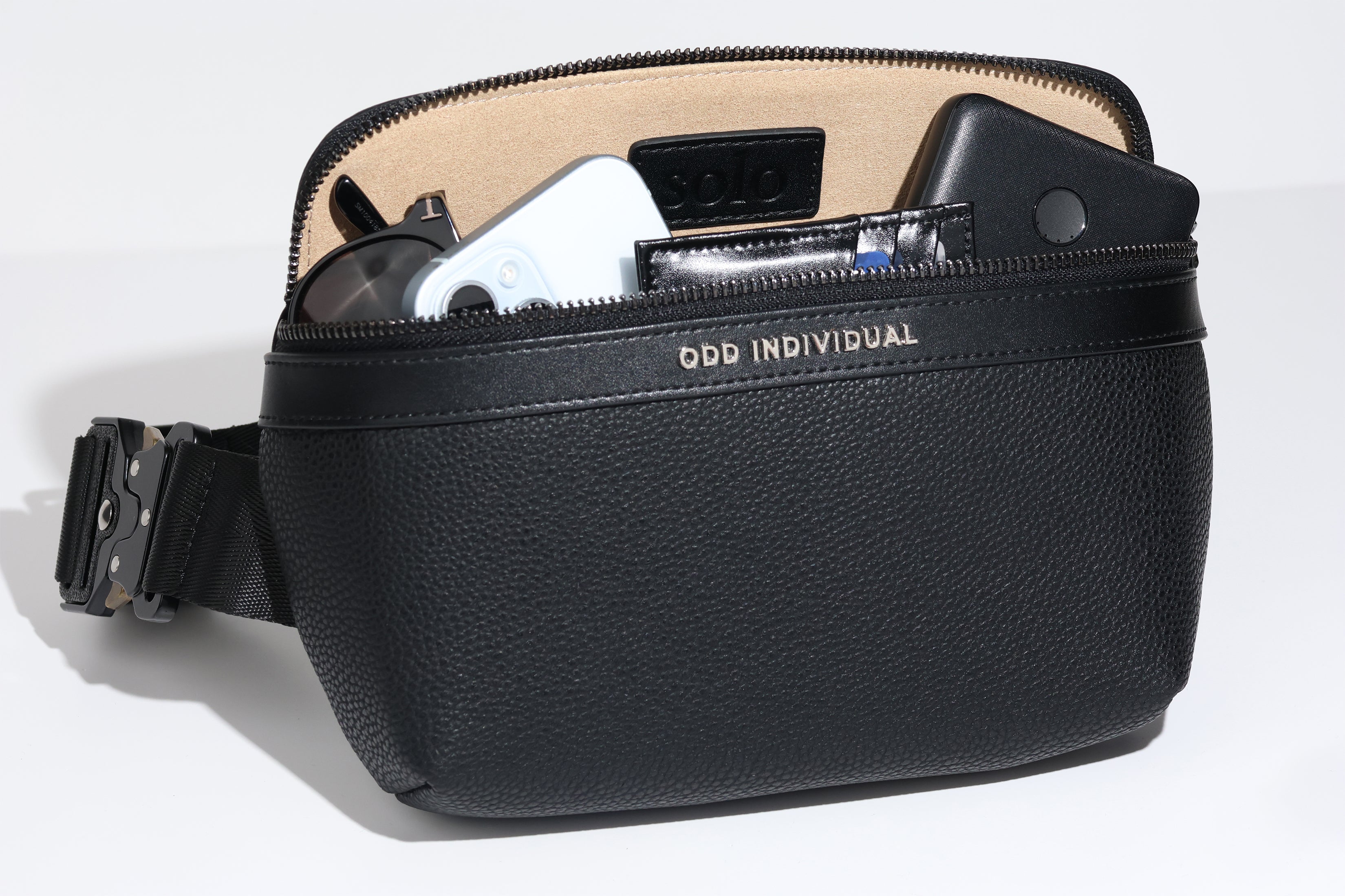Black leather sling bag/black leather belt bag/black leather crossbody bag/black leather waist bag/black leather travel pouch with brand name 'ODD INDIVIDUAL'  containing sunglasses, phone, wallet, and portable battery charger on a white background